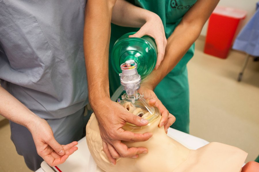 Two students with their arms entwined use a bag valve mask to provide manual ventilation to a patient.